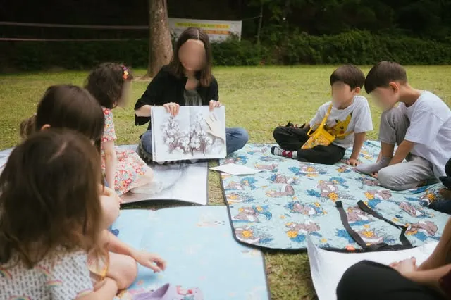 풀밭 위 돗자리에서 화가와 함께 그림을 그리는 아이들.